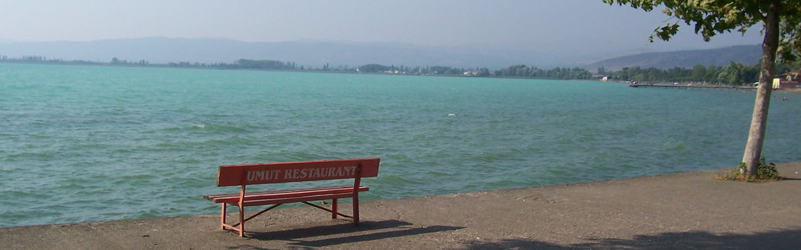 A red bench with the text "UMUT RESTAURANT" on the back. The bench is on a stone walkway overlooking a body of water, with buildings and large hills visible across the water.